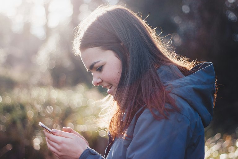 Junge Frau mit braunem langem Haar, die in beiden Händen ein Smartphone hält, in das sie lächelnd hineinschaut.