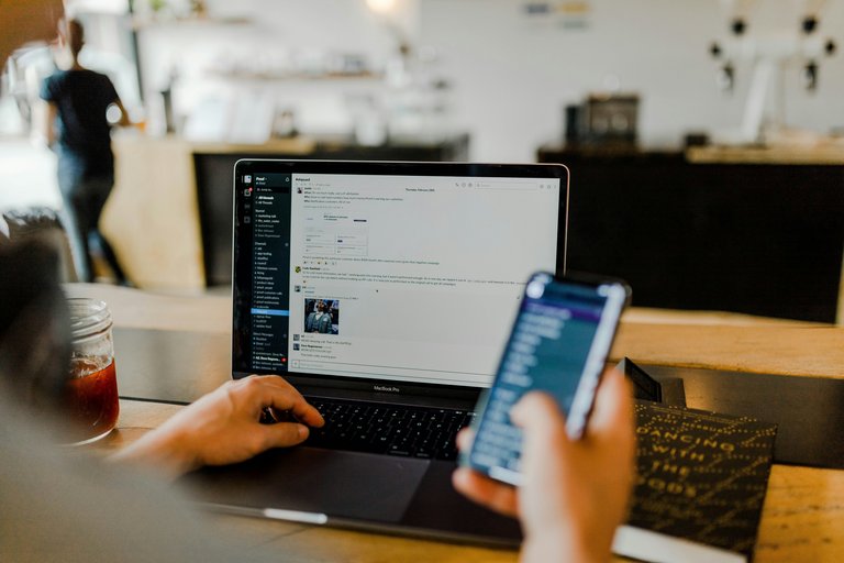 Mensch von hinten gesehen betrachtet Laptop-Bildschirm. Linke Hand liegt auf der Tastatur, während die rechte Hand ein Smartphone hält. 