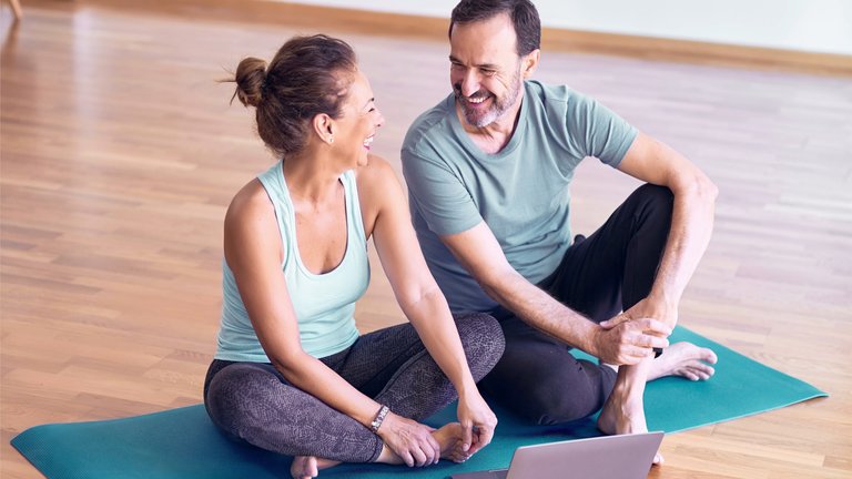 Ein lächelndes Paar mittleren Alters sitzt barfuß in Sportkleidung auf Yogamatten in einem hellen Raum und schaut sich gemeinsam etwas auf einem Laptop an.