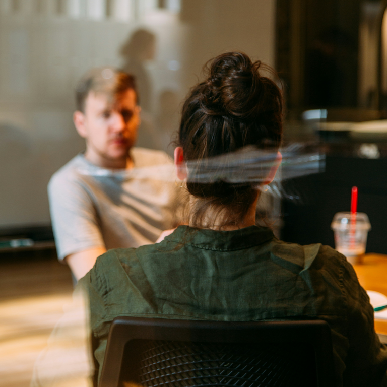Rückenansicht einer Frau im Gespräch mit einem Mann an einem Tisch; durch eine spiegelnde Glasscheibe fotografiert, wirkt die Szene diskret und vertraulich.