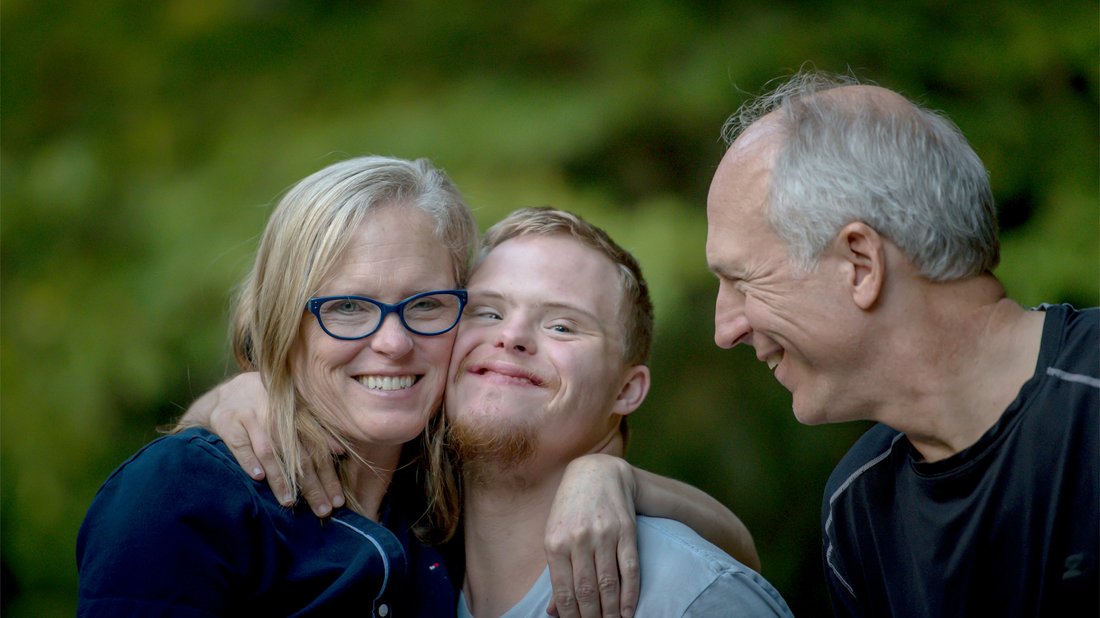 Drei Menschen stehen eng beieinander im Freien, lächeln und umarmen sich herzlich vor grünem Hintergrund.