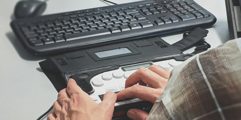 Person tippt auf einer Braillezeile mit großen weißen Tasten, vor einer herkömmlichen Computertastatur – assistive Technologie zur barrierefreien Computernutzung für sehbehinderte oder blinde Menschen.